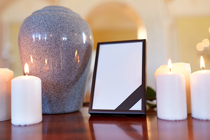 funeral and mourning concept - photo frame with black ribbon, cremation urn and burning candles on table in church