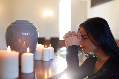 people and mourning concept - sad woman with cinerary urn and candles praying at funeral in church