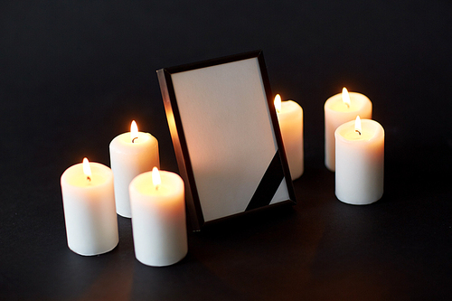 funeral and mourning concept - empty photo frame with ribbon and burning candles over black background