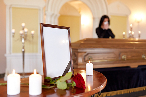 people and mourning concept - photo frame with black ribbon, candles and woman crying near coffin at funeral in church