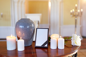 funeral and mourning concept - photo frame with black ribbon, cremation urn and burning candles on table in church