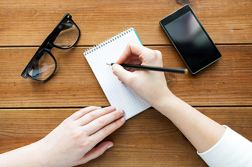 education, business and people concept - close up of woman or student writing to notepad with pencil and eyeglasses with smartphone on wooden table