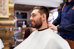 grooming, hairdressing and people concept - hairdresser or barber and man in cape with beard at barbershop