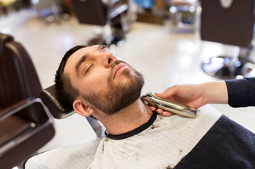 grooming and people concept - man and barber with trimmer or shaver cutting beard at barbershop