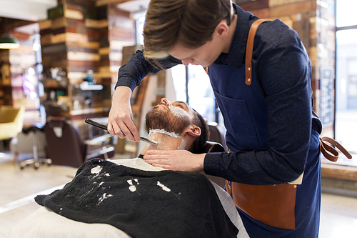 grooming and people concept - barber with straight razor shaving clients beard at barbershop