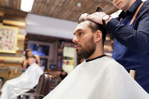 grooming, hairdressing and people concept - man and barber with comb and scissors cutting hair at barbershop