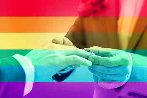 people, homosexuality, same-sex marriage and love concept - close up of happy male gay couple hands putting wedding ring on over  flag stripes background