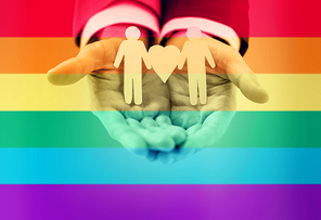 people, homosexuality, same-sex marriage and love concept - close up of happy male gay couple holding paper cutout love symbol over  flag stripes background