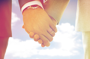 people, homosexuality, same-sex marriage and love concept - close up of happy male gay couple holding hands with wedding rings on over sky and sun background