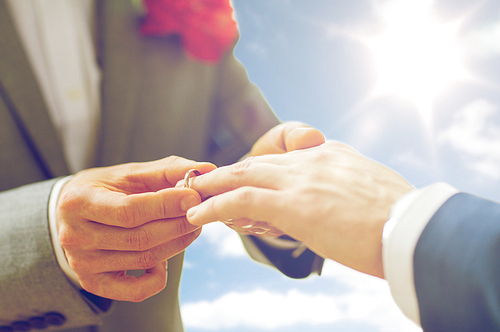 people, homosexuality, same-sex marriage and love concept - close up of happy male gay couple hands putting wedding ring on over sky and sun background
