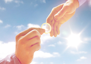 people, homosexuality, safe sex, sexual education and charity concept - close up of happy male gay couple hands giving condom over sky and sun background