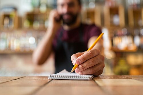 small business, people and service concept - bartender with notebook and pencil calling on phone at bar or coffee shop