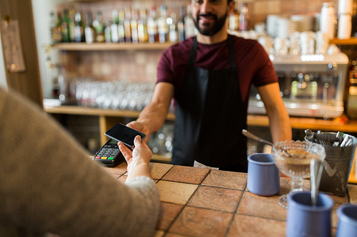 modern technology and people concept - man or bartender with payment terminal and customer hand with smartphone at bar of coffee shop