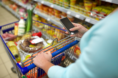 consumerism, technology and people concept - woman with smartphone and shopping cart or trolley buying food at grocery store or supermarket