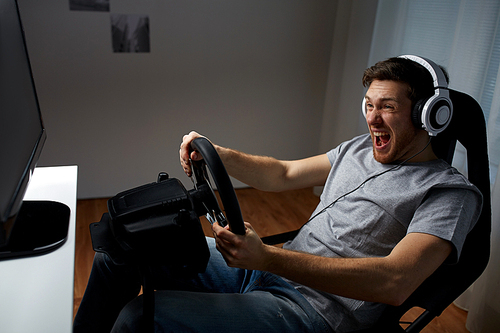 technology, gaming, entertainment and people concept - young man in headphones with pc computer playing car racing video game at home and steering wheel
