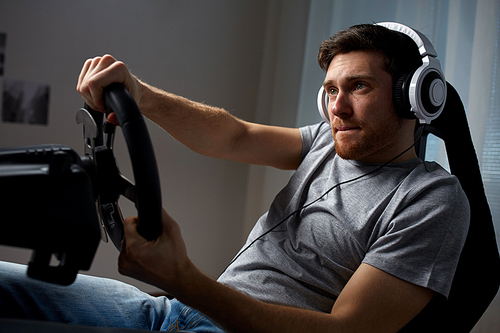 technology, gaming, entertainment and people concept - young man in headphones with pc computer playing car racing video game at home and steering wheel