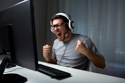 technology, gaming, entertainment, let's play and people concept - happy young man in eyeglasses with headset playing and winning computer game at home and streaming playthrough or walkthrough video