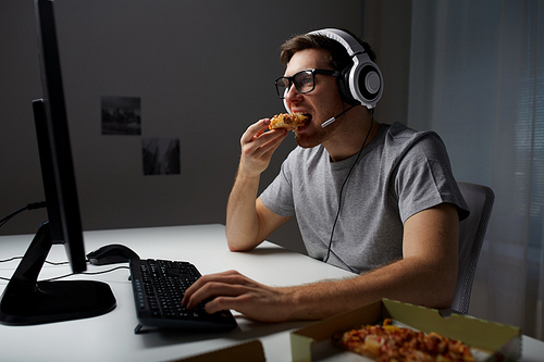 technology, gaming, entertainment, let's play and people concept - young man in headset with pc computer eating pizza while playing game at home and streaming playthrough or walkthrough video