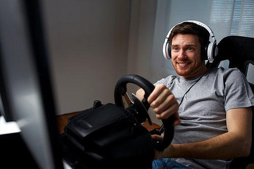 technology, gaming, entertainment and people concept - happy smiling young man in headphones with pc computer playing car racing video game at home and steering wheel