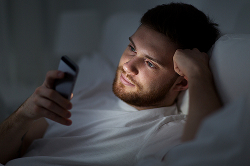 technology, internet, communication and people concept - young man texting on smartphone in bed at home at night