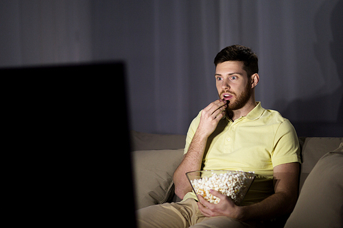 people, mass media, television and entertainment concept - young man watching tv and eating popcorn at night at home