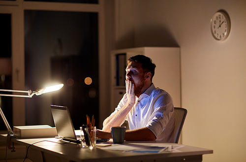 business, overwork, deadline and people concept - tired man with laptop working at night office and yawning
