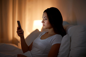 technology, internet, communication and people concept - young woman with smartphone in bed at home at night