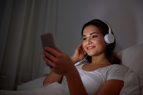 technology, leisure and people concept - happy smiling young woman with smartphone and headphones listening to music in bed at night