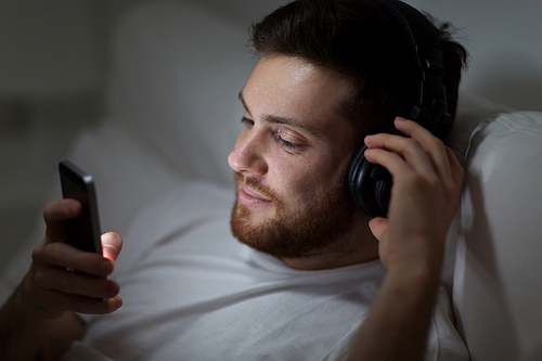 technology, internet, communication and people concept - happy smiling young man with smartphone and headphones hones listening to music in bed at night