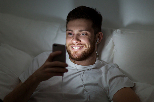technology, internet, communication and people concept - happy smiling young man with smartphone and earphones listening to music in bed at night