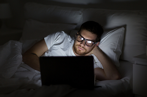 technology, internet, communication and people concept - young man in glasses with laptop computer in bed at home bedroom at night