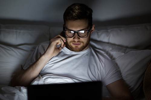 technology, internet, communication and people concept - young man in glasses with laptop computer calling on smartphone in bed at home bedroom at night
