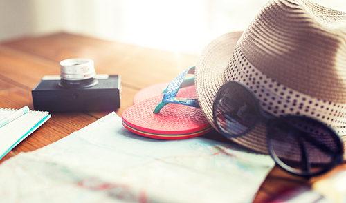 summer vacation, tourism and objects concept - close up of travel map, flip-flops, hat and camera on wooden table at home