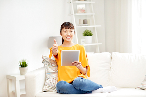 people, technology, gesture and leisure concept - happy young asian woman sitting on sofa with tablet pc computer showing thumbs up at home