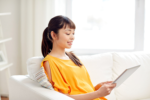 people, technology and leisure concept - happy young asian woman sitting on sofa with tablet pc computer at home