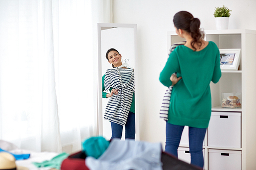 tourism, people and luggage concept - happy young woman packing travel bag at home or hotel room and looking to mirror