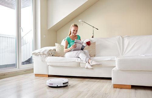 people, housework and technology concept - happy woman reading magazine and drinking coffee while robot vacuum cleaner at home