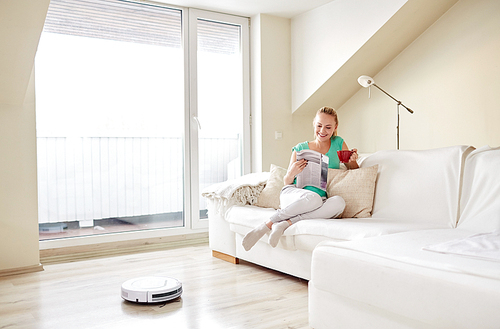 people, housework and technology concept - happy woman reading magazine and drinking coffee while robot vacuum cleaner at home