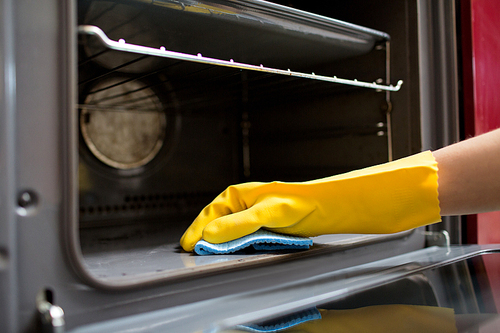 people, housework and housekeeping concept - hand in rubber glove with rag cleaning oven at home kitchen