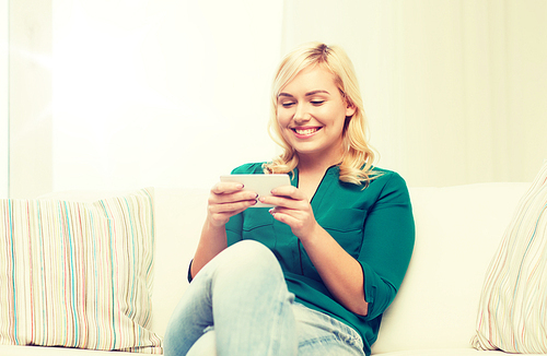 people, technology, communication and leisure concept - happy young woman sitting on sofa with smartphone at home