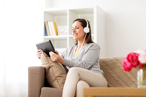 people, technology and leisure concept - happy woman sitting on sofa with tablet pc computer and headphones listening to music at home