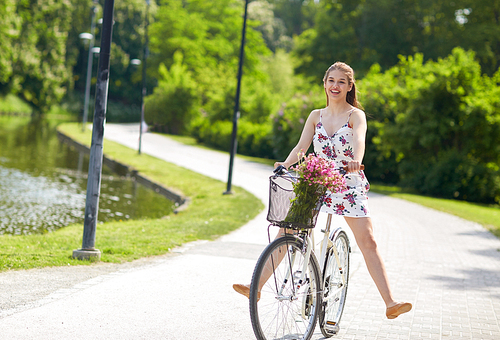 people, leisure and lifestyle - happy young hipster woman wearing summer dress riding fixie bicycle with wild flowers in basket at park