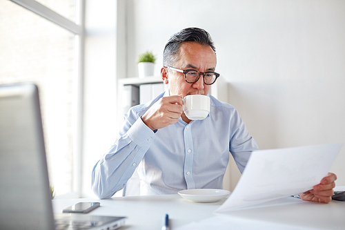 business, people, paperwork and technology concept - businessman reading paper and drinking coffee at office