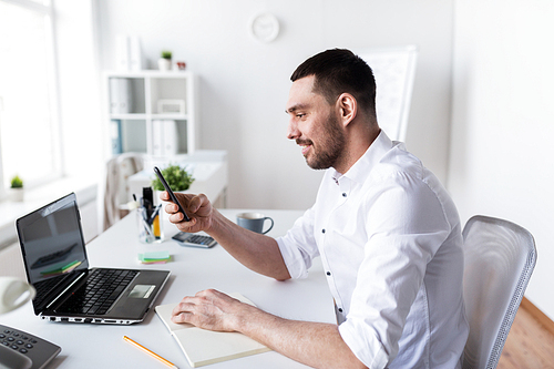 business, people and technology concept - businessman with smartphone, laptop computer and notebook at office