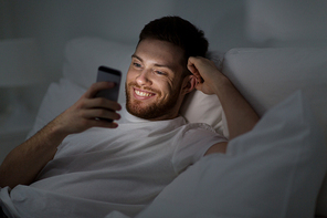 technology, internet, communication and people concept - happy smiling young man texting on smartphone in bed at home at night