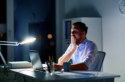 business, overwork, deadline and people concept - tired man with laptop working at night office and yawning