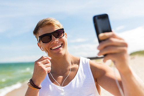 summer holidays and people concept - happy smiling young man with earphones and smartphone taking selfie on beach