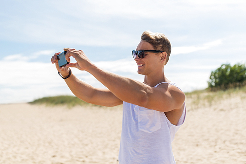 summer holidays and people concept - happy smiling young man with smartphone on beach photographing