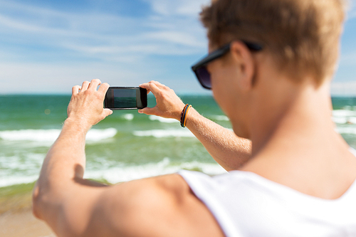summer holidays and people concept - young man with smartphone on beach photographing sea