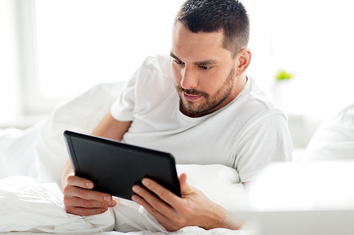 technology, internet, communication and people concept - young man with tablet pc computer in bed at home bedroom